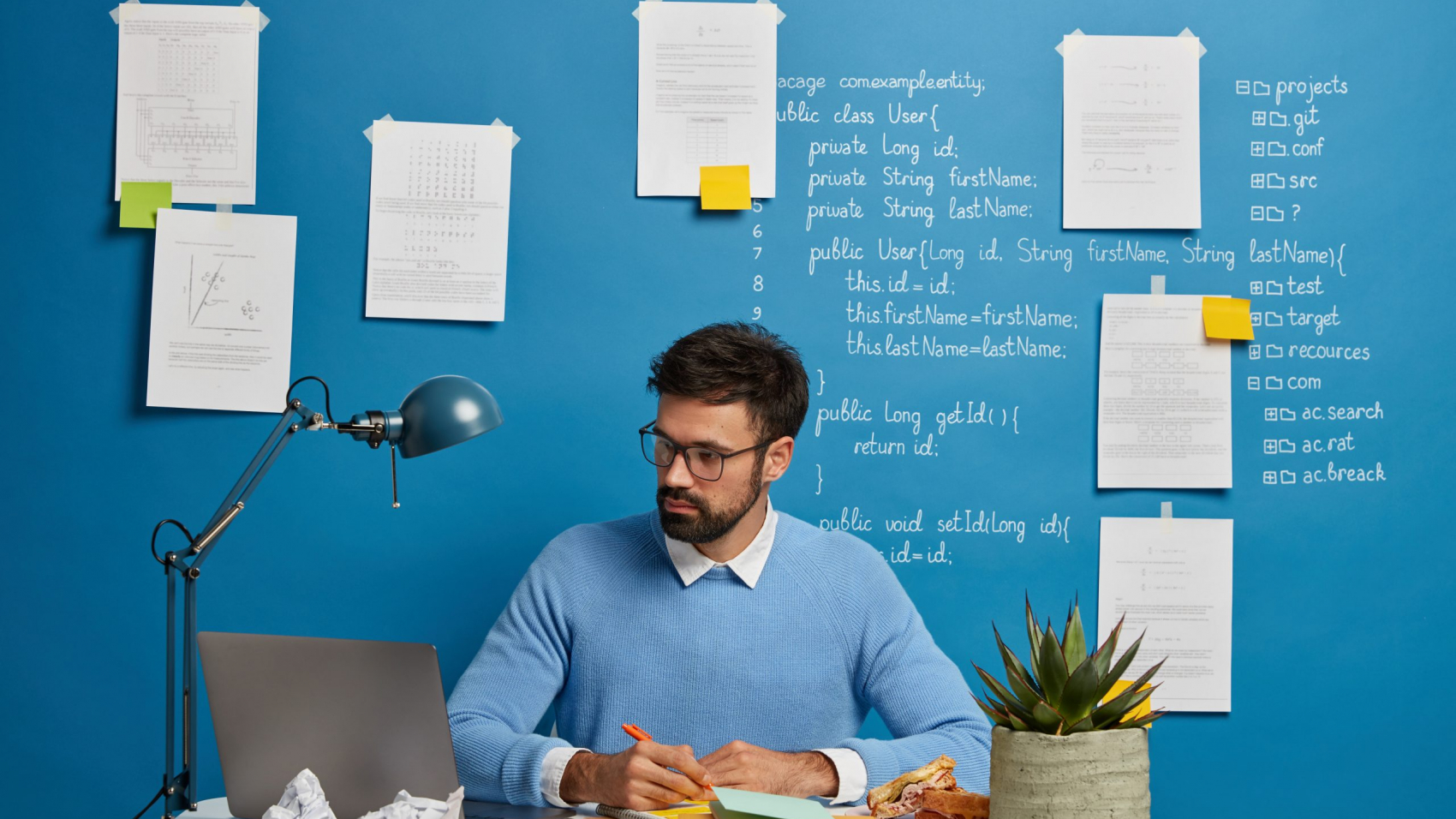 Young millennial student searches information for homework, writes down necessary content in textbook, prepare project on IT topic, sits at desktop with laptop computer, stuck papers on blue wall