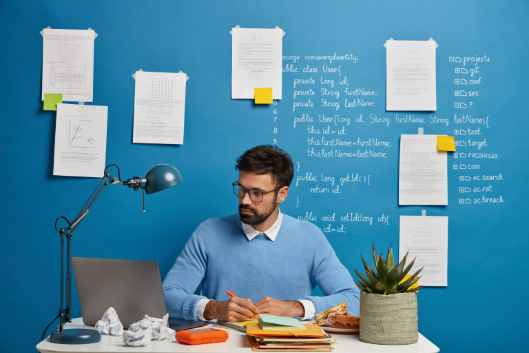Young millennial student searches information for homework, writes down necessary content in textbook, prepare project on IT topic, sits at desktop with laptop computer, stuck papers on blue wall